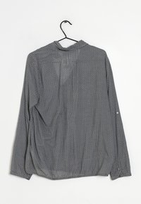 Grau, langärmliges Hemd mit weißem geometrischem Muster, strukturiertem Stoff, Knopfakzenten an den Ärmeln und einem abgerundeten Saum. Hängt an einem schwarzen Haken.