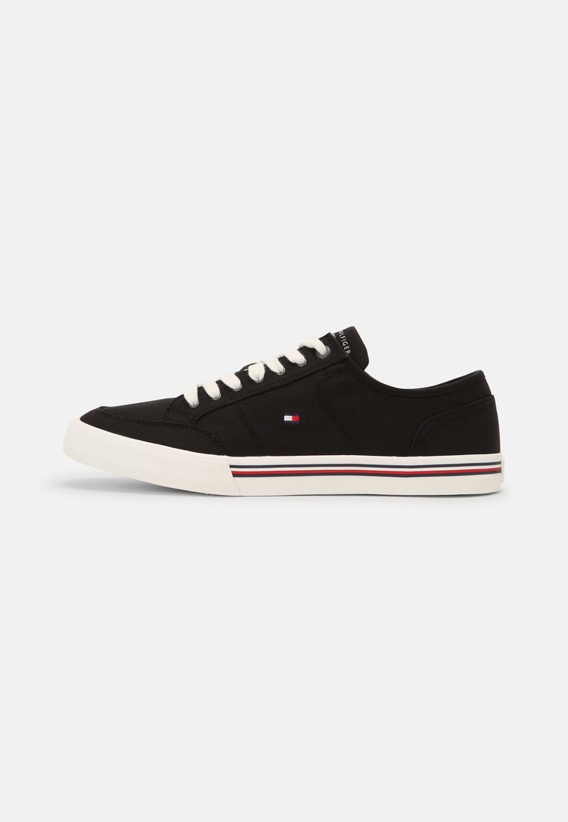 Baskets basses noires avec semelle et lacets blancs, avec un petit logo rouge, blanc et bleu sur le côté et un détail rayé sur la semelle.