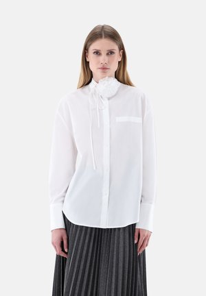 Woman wearing a white button-up shirt with a large fabric flower tied at the collar and a dark gray pleated skirt.