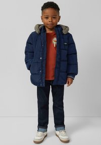 Marineblauwe gewatteerde jas met een grijze fleece-voering in de capuchon, een rode shirt met grafisch ontwerp, donkere jeans en witte schoenen met lichte zolen.
