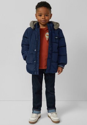 Doudoune bleu marine avec une capuche en polaire grise, chemise rouge avec un design graphique, jeans foncés, et chaussures blanches avec semelles claires.