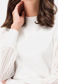 Femme aux cheveux bruns mi-longs portant un pull blanc avec des manches rayées transparentes, touchant son cou avec sa main gauche.