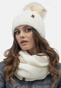 Gorro de lana blanco tejido con un pom-pom esponjoso en la parte superior, acompañado de una bufanda infinity texturizada a juego; ambos presentan un sutil acento de cuero marrón.