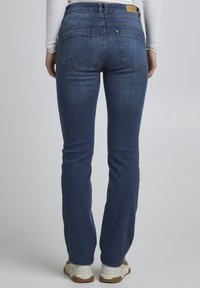 Tamsiai mėlyni džinsai su vidutinio aukščio juosmeniu, dviem užpakalinėmis kišenėmis ir subtiliais siuvinėjimo motyvais, derinami su šviesios spalvos sportinėmis avalynėmis.