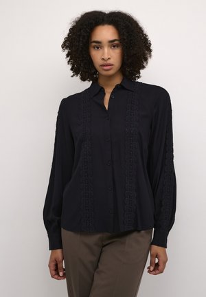 Blouse marine à manches longues, avec un devant boutonné, des accents de dentelle détaillés le long des côtés et une coupe décontractée. Texture de tissu lisse.