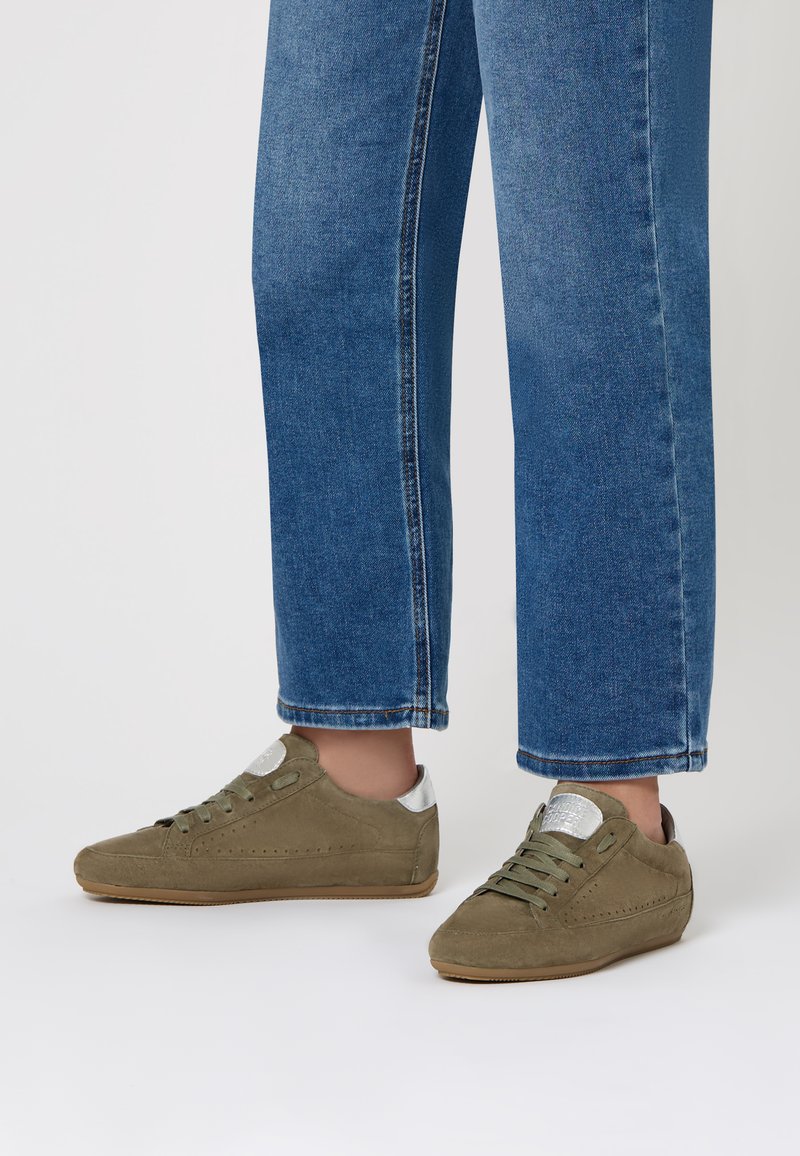 Sneakers in suede verde oliva con dettagli in argento, punta tonda e decorazioni perforate, abbinati a jeans blu a vita alta.