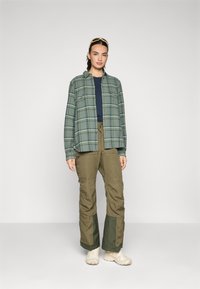 Grön rutad skjorta över en mörkblå topp, ihop med olivgröna cargobyxor. Ljusfärgade sneakers fullbordar outfiten. Tydliga hantverksdetaljer synliga.