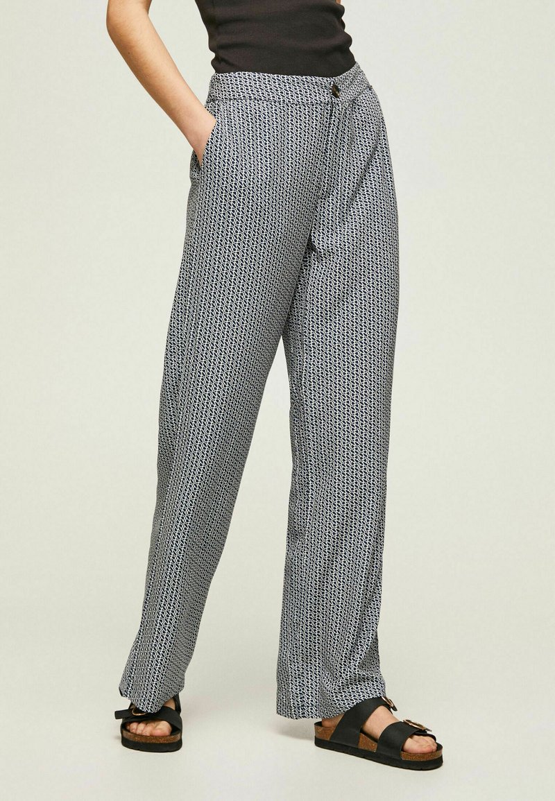 Pantaloni leggeri a fantasia in bianco e nero con vita alta, tasche laterali e chiusura a bottone, abbinati a sandali neri.