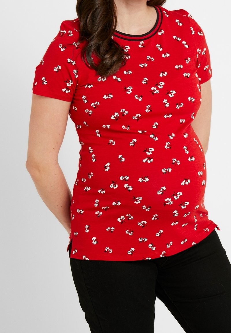 Röd kortärmad graviditetstopp med vit och svart blommönster, rund halsringning med svart kant, och sprund på sidorna vid fållen.