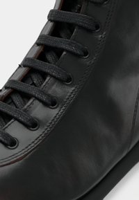 Gros plan d'une chaussure en cuir noir avec des lacets noirs texturés passés à travers six paires d'œillets, montrant une finition lisse et des détails de couture.