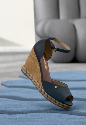 Námornícky modrá klinová sandála so zapínaním okolo členka, korkovým platformovým opätkom a otvorenou špičkou na textúrovanom modrom povrchu s rozmazaným sivým pozadím.