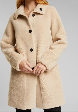 Beige fleecejacka med krage, knapprad fram och sidofickor. Utmärkande texturkontrast på ärmar och fåll. Svarta knappar ger detaljer.