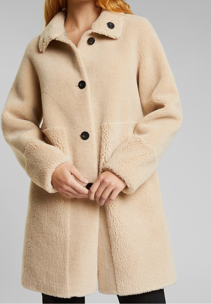 Beige fleecejacka med krage, knapprad fram och sidofickor. Utmärkande texturkontrast på ärmar och fåll. Svarta knappar ger detaljer.