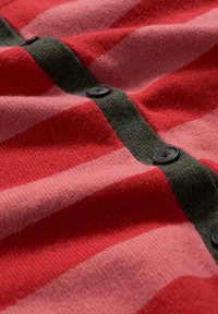 Stickad kofta med horisontella ränder i röd och ljusrosa, med en mörkgrön knappslå och svarta knappar. Mjuk textur.