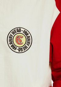 Czerwona i biała koszula z długim rękawem z okrągłym haftowanym wzorem z tekstem oraz stylizowaną grafiką na środku.