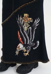 Zwarte denim rok met een rafelrand, voorzien van geborduurde creaties van een adelaar, kruis en rozen in levendige kleuren. Gecombineerd met zwarte laarzen.