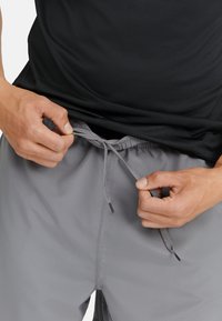 Herren grau Shorts mit einem elastischen Bund und Kordelzug. Das Oberteil ist ein schwarzes, strukturiertes Tanktop. Hände, die den Bund anpassen.