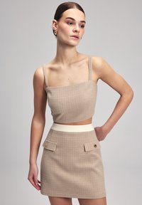 Beige geruite crop top met dunne bandjes, gecombineerd met een beige minirok met een contrasterende witte tailleband en twee voorzakken.
