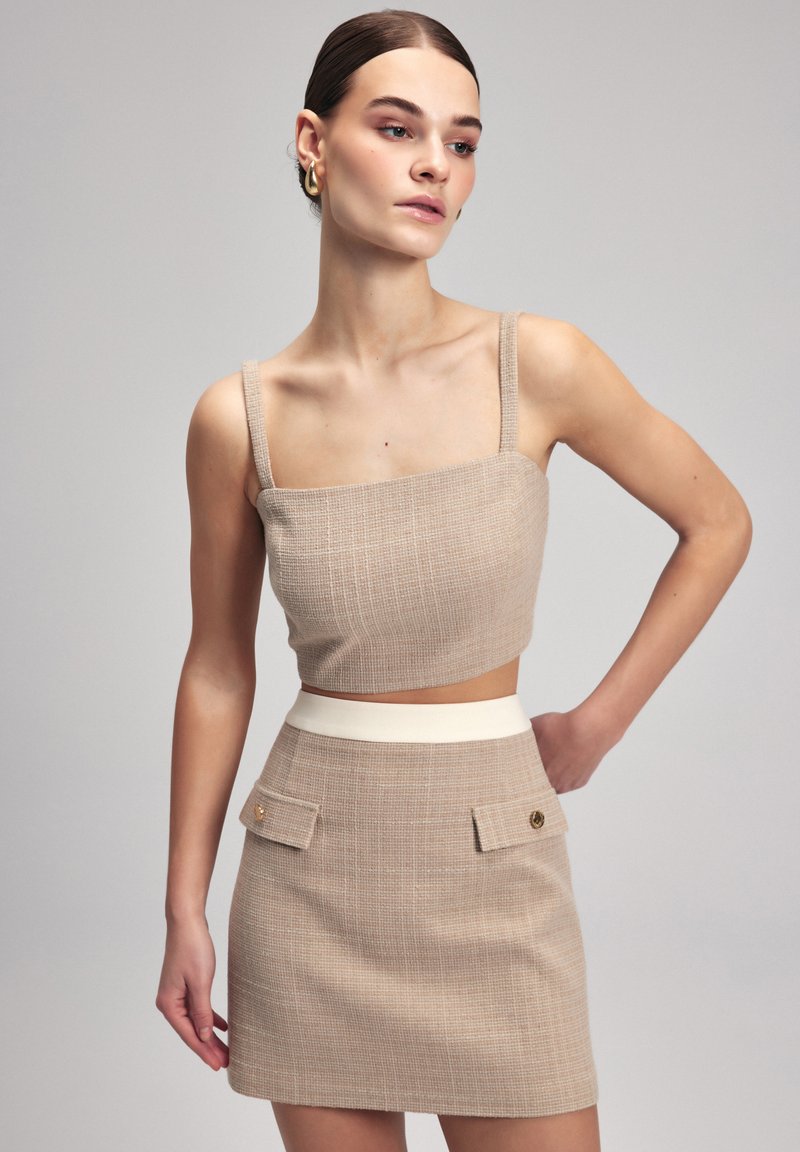Beige geruite crop top met dunne bandjes, gecombineerd met een beige minirok met een contrasterende witte tailleband en twee voorzakken.