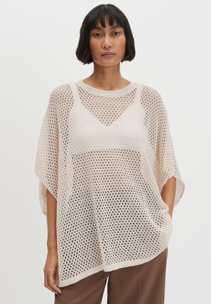 Frau mit kurzen schwarzen Haaren, die ein lockeres, cremefarbenes, offen gestricktes Oberteil über einem beigen Top und braunen Hosen trägt und vor einem schlichten Hintergrund steht.