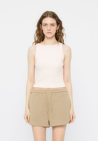 Νέα γυναίκα με κυματιστά μαλλιά, που φοράει λευκό crop top χωρίς μανίκια και μπεζ σορτς με κορδόνι, στέκεται μπροστά σε μια απλή λευκή επιφάνεια.