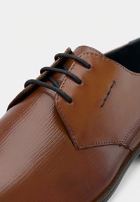 Scarpa da cerimonia in pelle marrone con lacci neri e superficie lucida e texturizzata, vista da vicino con un focus sulla parte superiore anteriore.