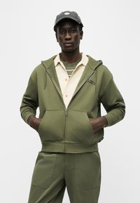 Νέος άντρας που φοράει φούτερ με φερμουάρ σε ελιά πράσινο χρώμα, παντελόνι στο ίδιο χρώμα, κρεμ πουκάμισο με κουμπιά, ριγέ μπλουζάκι και σκούρο γκρι καπέλο, στέκεται με τα χέρια στις τσέπες.