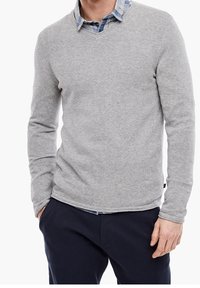 Homme portant un pull à col en V gris par-dessus une chemise à carreaux bleue avec un pantalon bleu marine foncé, les mains dans les poches sur fond blanc.