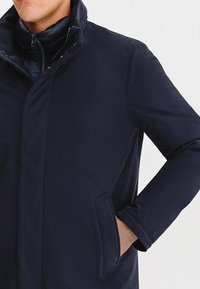 Granatowy płaszcz z wysokim kołnierzem, zapinany na zamek błyskawiczny, z bocznymi kieszeniami. Materiał jest gładki z lekko teksturowanym wykończeniem.