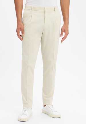 Man wearing beige slim-fit trousers and white sneakers, standing against a plain light background.