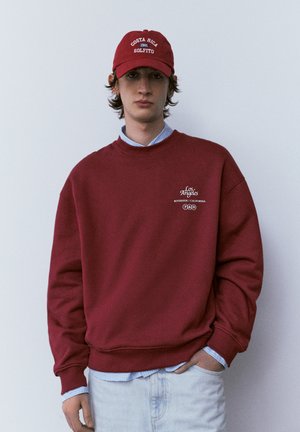 Mladi moški, oblečen v rdečo majico z napisom "Los Angeles Riverside California" in rdečo kapo "Costa Rica Golfito 1981", stoji ob navadni steni.