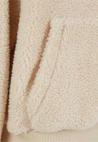 Giacca in pile beige con una superficie morbida e testurizzata, dotata di una tasca e polsini a costine all'orlo. Vista ravvicinata del dettaglio del tessuto.