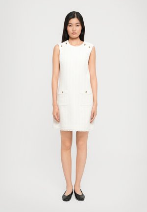 Abito bianco senza maniche con tessuto texturizzato, due tasche frontali e bottoni dorati sulle spalle; silhouette dritta con orlo sfrangiato.