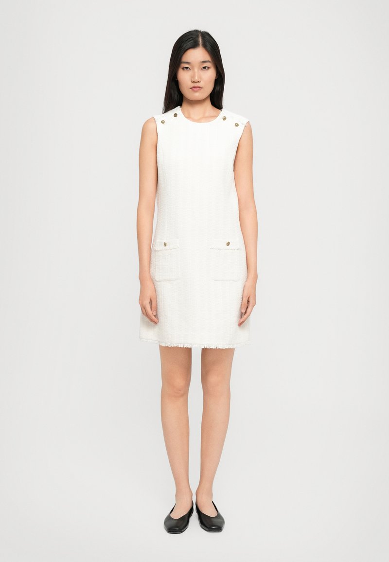 Biała sukienka bez rękawów z teksturowanej tkaniny, z dwiema przednimi kieszeniami i złotymi guzikami na ramionach; prosty fason z postrzępionym dołem.
