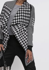 Svetr s černo-bílým vzorem, obsahující houndstooth, pruhy a kostky. Měkký pletený materiál s volně drapovaným předním dílem a dlouhými rukávy.