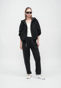 Svart hettegenser med glidelås og store frontlommer, sammen med svarte joggebukser med en liten logo. Brukt med hvite slip-on sneakers og solbriller.