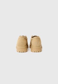 Tan suede shoes with a thick, textured sole, featuring a rounded design and logo detail on the back. Simple and understated craftsmanship.