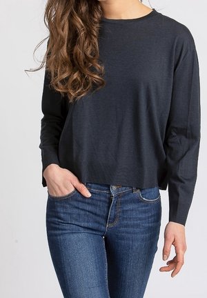 Kobieta z długimi falowanymi brązowymi włosami, ubrana w ciemnoszary sweter z długim rękawem i niebieskie dżinsy, z jedną ręką w kieszeni, stojąca na jednolitym tle.