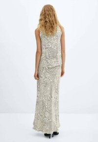 Maxikjole i lys beige med paisleymønster. Ærmeløs design, flydende tekstur og en let slæb ved kanten. Sorte høje hæle synlige.