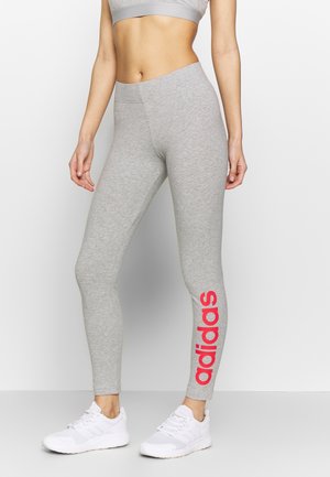 Leggings grigi con un design aderente, caratterizzati da un grande logo rosso "adidas" sulla gamba sinistra. Realizzati in tessuto elastico, abbinati a sneakers bianche.