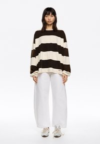 Gestreepte oversized trui in crème en donkerbruin, met lange mouwen. In combinatie met witte broeken met wijde pijpen en zilveren sneakers.
