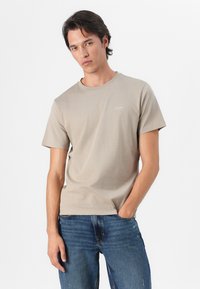 Beige short-sleeve t-shirt with a round neckline and subtle brand logo printed on the chest, worn with fitted blue jeans.