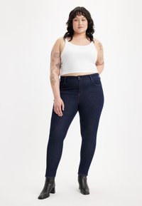 Jeans ajustados de color azul oscuro con una cintura alta, hechos de denim, combinados con una camiseta sin mangas ajustada blanca. Botines negros de tacón completan el look.