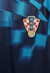 Maillot de sport bleu et noir avec un motif dégradé ; présente un blason brodé avec des carreaux rouges et blancs et du texte bleu.