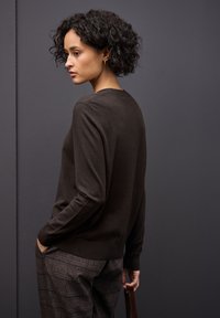 Brązowy sweter dzianinowy z długimi rękawami, okrągłym dekoltem i ribbowanym dołem, zestawiony z szarymi spodniami w kratę. Modelka ma kręcone włosy.