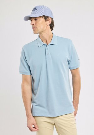 Homme portant un polo bleu clair et une casquette rayée bleue, regardant vers la gauche sur un fond uni.