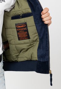 Olijfgroene bomberjack met marineblauwe corduroy accenten, gewatteerde voering en oranje label, voorzien van een ritsluiting en een binnenzak.