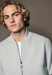 Light grey zip-up bomber jacket made of textured fabric, featuring a ribbed collar and metal zipper. Plain white t-shirt underneath.