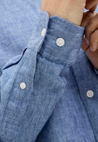 Puño de camisa de lino azul claro con dos botones blancos y textura visible; presenta un collar abotonado y un ajuste ligeramente relajado.