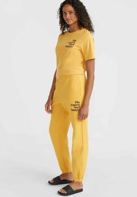 Gul matchande set bestående av en kortärmad T-shirt med svart text och sweatpants med elastiska muddar och fickor, kompletterat med svarta sandaler.